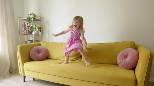 Cheerful and Cute Preschool Girl Jumps and Dances on Yellow Large Sofa in Living Room