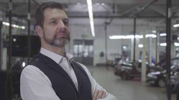 Close-up of Thoughtful Caucasian Man with Beard Standing at the Background of Cars in Repair Shop