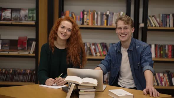 Two Young Male and Female European Students Sitting at the Table with Books and Laptop in the