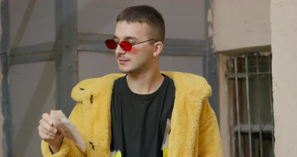 Stylish and Daring Young Man in Glasses with Red Lenses and Yellow Faux Fur Coat He Stands at Ease