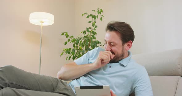 Young Bearded Man is Lying on the Sofa and Holding Tablet in Hands and Suddenly Sneezes White Walls