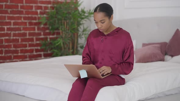 Young Intelligent Concentrated Woman Typing on Laptop Keyboard Sitting on Bed in the Morning