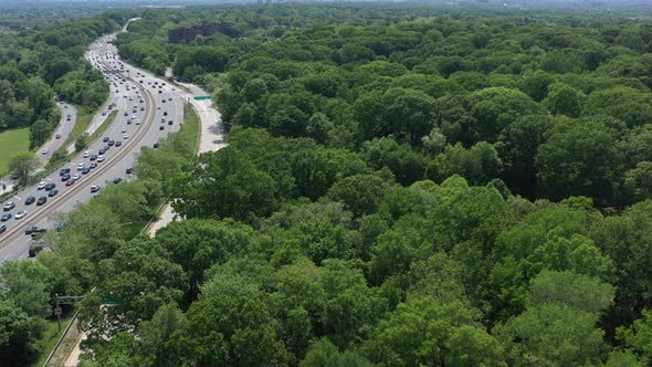 An aerial view over a busy parkway next to a large park with green trees. The drone camera dolly in