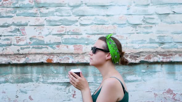 Young woman with bandana on hair standing on turquoise brick wall background with coffee to go city