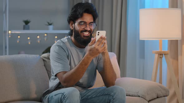 Happy Arabian Indian Male Millennial Man Holding Modern Smartphone Texting Message at Home Sitting