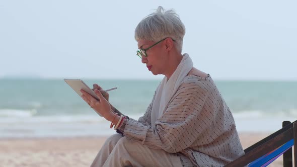 Asian elderly woman using a tablet computer while sitting on the beach.