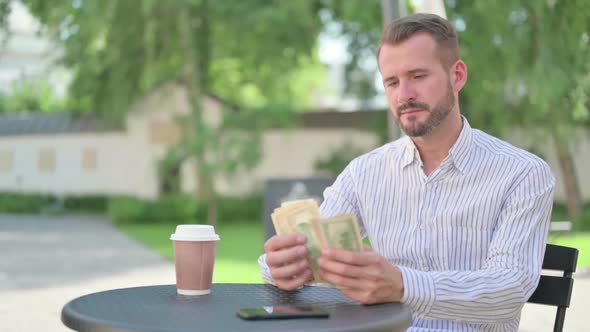 Middle Aged Man Feeling Happy While Counting Dollars in Outdoor Cafe