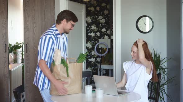 Happy Family in Kitchen, Husband Brings a Lot of Useful Food and Wife Works Behind Laptop