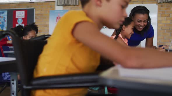 Mixed race schoolboy in wheelchair, sitting in classroom making notes, teacher in background