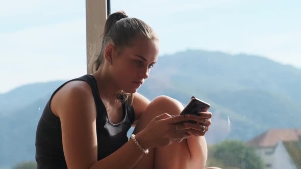 Pretty Young Woman with Smartphone Sits on the Windowsill By the Window in City