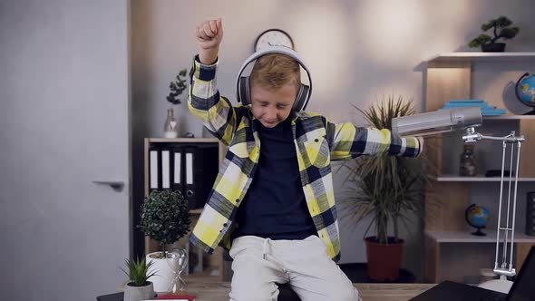 Happy Boy in Stylish Clothes Sitting on Knees on His Table and Enjoying Music in Headphones