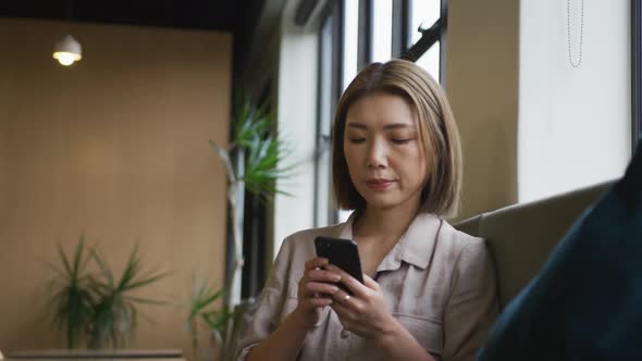 Asian businesswoman sitting by a window using smartphone in modern office