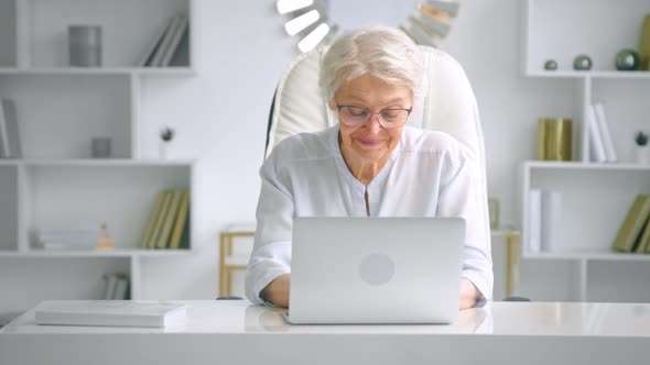 Aged businesswoman with glasses types on grey laptop with cheerful smile