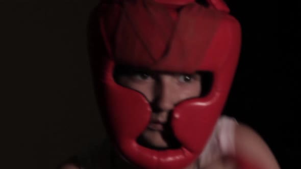 Young guy boxer practices punches in a dark room. Boxing protective helmet and hand bandages