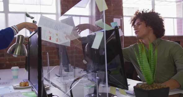 Diverse male and female business colleagues sitting at desks passing document over sneeze guard