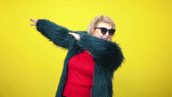 Stylish Senior Caucasian Woman in Fur Coat and Sunglasses Stretching Hands Aside at Yellow