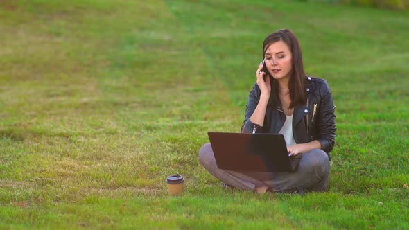 A Girl in the Park on the Lawn Working on a Laptop, Phone Rings, Answers an Incoming Call.