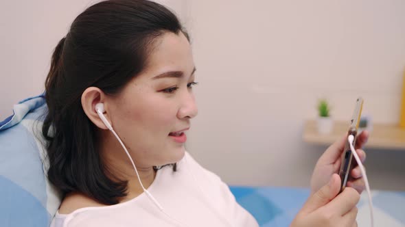 Cheerful young beautiful Asian woman relaxing and listening to music by tablet with headphones on th