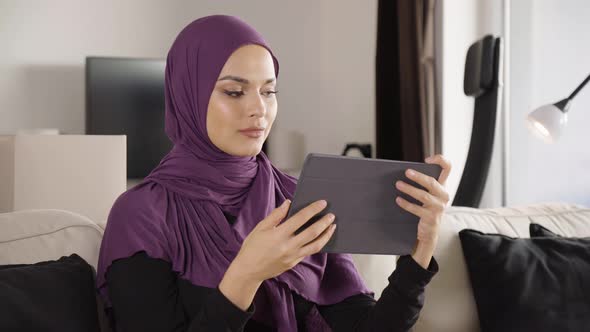 A Young Beautiful Muslim Woman Works on a Tablet with a Smile As She Sits on a Sofa in an Apartment