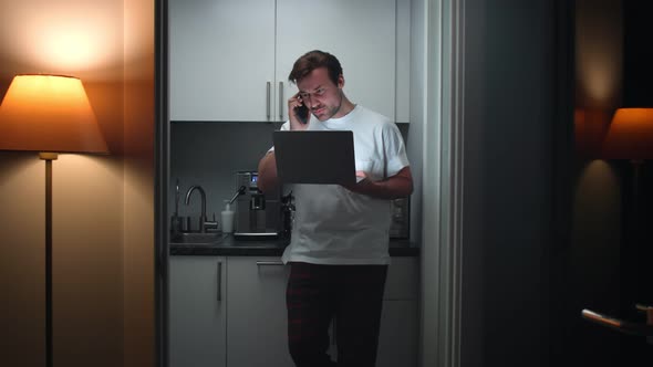 Man in Pajamas Stand in Kitchen Talking on Smartphone and Working on Laptop