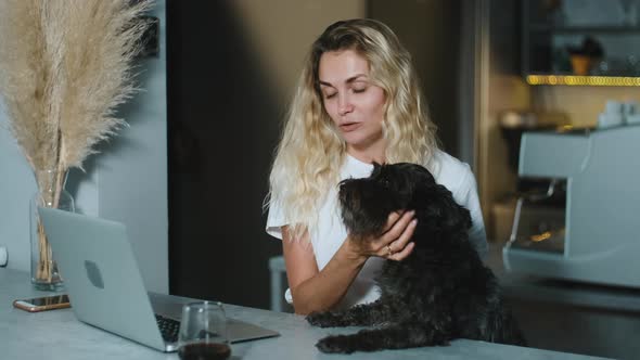 Happy smiling blonde woman hugging adorable puppy dog and looking qt laptop working at coffee shop.