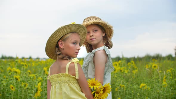 Two Teen Girls in Straw Hat and Summer Dress Posing on Sunflowers Field Landscape in Countryside