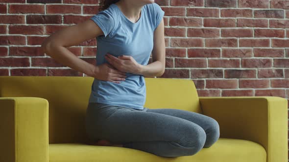 Woman is sitting on sofa and clutching her side with hands from pain in liver. Woman with spasms