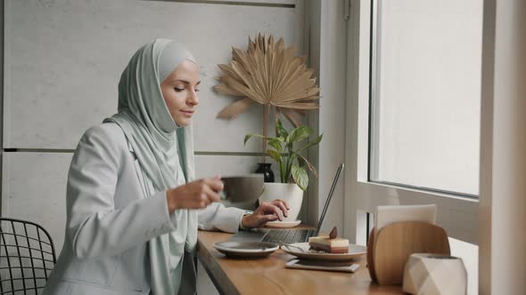 Arab Girl Freelancer Working with Laptop and Drinking Coffee with Dessert Indoors in Cafe