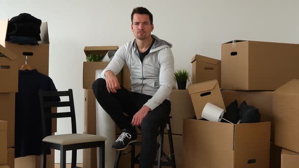 A Moving Man Sits on a Chair and Looks Seriously at the Camera in an Empty Apartment