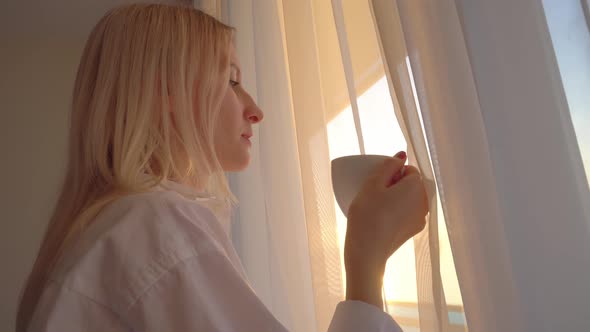 blonde in a white shirt looks out window, drinks a hot drink of tea or coffee,