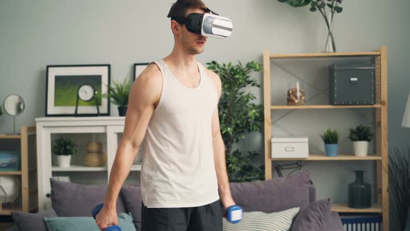 Young Man in Trendy Virtual Reality Glasses Is Exercising with Dumbbells at Home