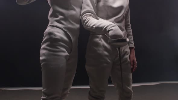 Two Young Women Fencers in White Protective Suits Standing in the Dark Studio