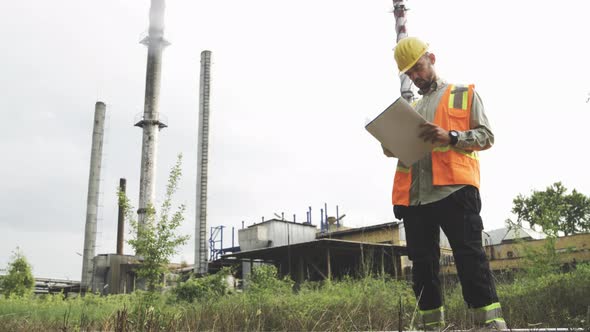 Male Architect Reviewing Blueprint at Construction Site Against Tall Chimneys