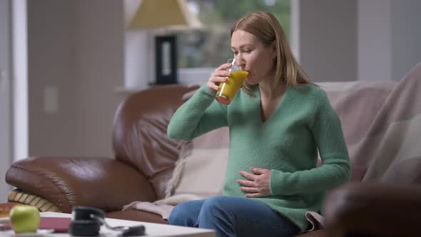 Happy Young Pregnant Woman Drinking Healthful Orange Juice Caressing Belly in Slow Motion Sitting on