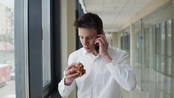 Young Man in White Shirt Talks on the Mobile Phone and Eats the Burger Indoors