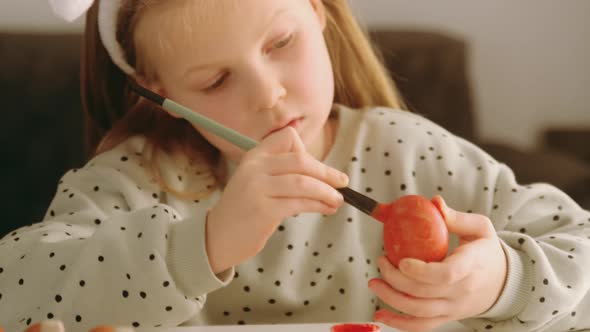 Little child girl with rabbit ears decorates Easter eggs with colored food paint.