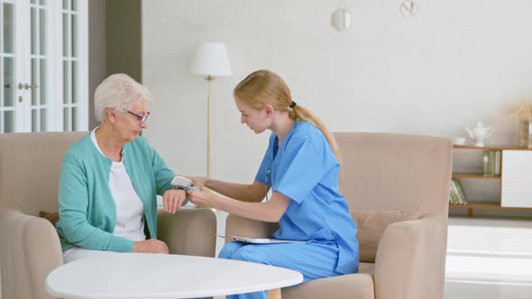 Skilled nurse in blue uniform puts digital blood pressure monitor on senior woman hand