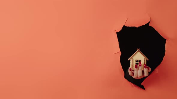 Close Up of Woman's Hand Holding Small Wooden House Through a Hole in the Orange Wall in the Studio