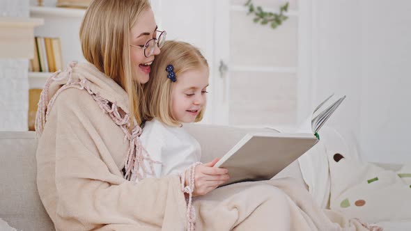 Young Caring Caucasian Mother Mum Blonde Woman Sits with Daughter on Couch Reading Book Literature