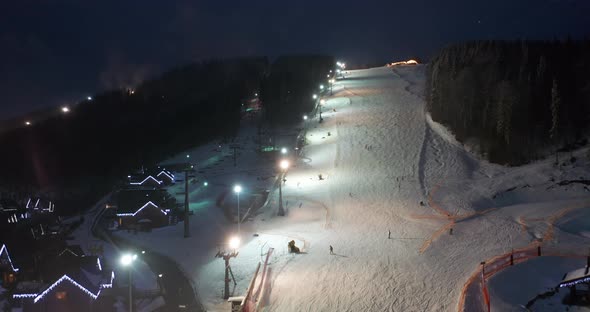 Aerial View of Skiers Go Down Ski Slopes Near Ski Lifts on Ski Resort at Night.