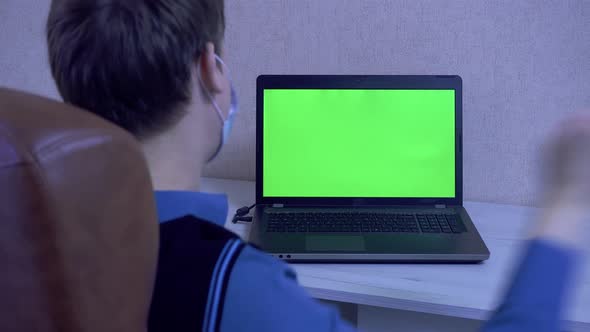 A Man Dances at a Computer at a Videoconference in a Protective Face Mask a Chroma Key on a Laptop