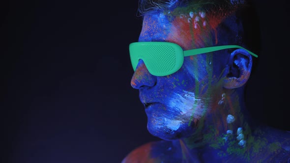 A Man with UV Drawings and Green Neon Glasses Inflates a Bubble of Bubblegum