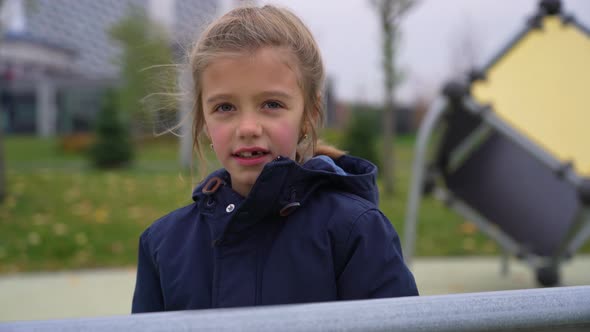 Little Girl Putting on Knitted Hat on Head and Looking in Camera Standing in Playground Autumn Park