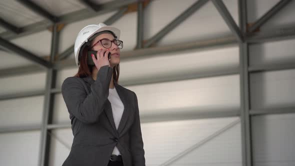 A Young Woman in a Helmet Speaks on the Phone at a Construction Site. The Boss in a Suit Is Talking