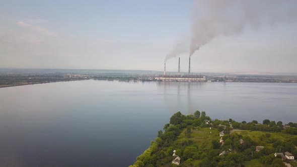 Aerial view of small village between green trees and big lake with high chimney pipes polluting 