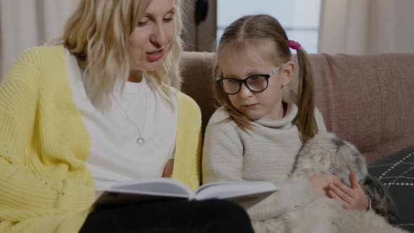 Mom and Daughter Reading Book and Sitting on Couch with Dog in Home Room Rbbro