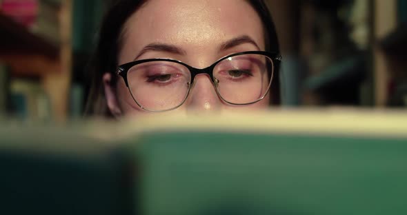 Close Portrait of Girl in Glasses Reading Book Leads Eyes at Camera in Library