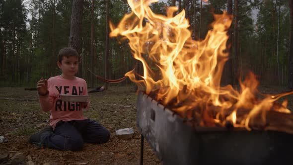 Bright Flame From Barbecue Grill Illuminate Silhouette of Boy Sitting Near Fire