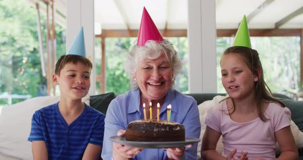 Caucasian senior woman in party hat blowing candles on birthday cake while her grandchildren sitting