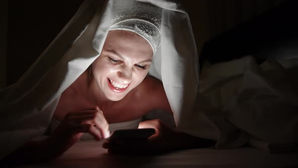 Beautiful Young Woman Using Smart Phone Under Blanket in Bedroom on Bedtime